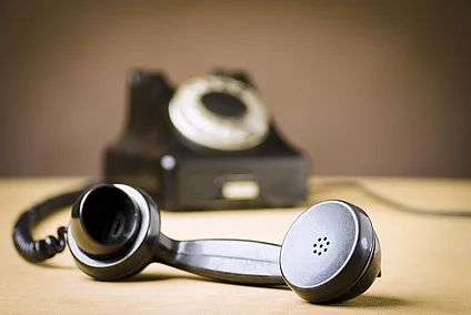 A vintage black rotary telephone with its handset resting off the hook in the foreground, placed on a wooden surface, with the main body of the phone in the background.