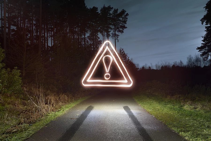 A glowing triangular warning sign with an exclamation mark above a dark, empty road at night, flanked by trees on either side.
